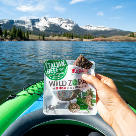 A hand holds a Wild Zora Bars - Italian Beef Bar With Garlic, Oregano & Basil 10-Pack above a kayak on a lake, framed by pine trees and snowy mountains under a blue sky. Enjoy this AIP-friendly protein bar in the heart of nature.