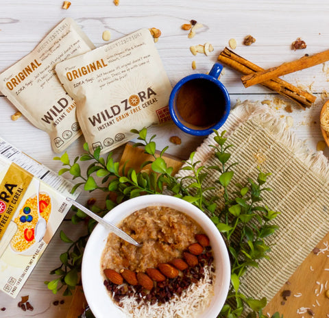 A bowl of Wild Zora Instant Cereal - Original With Almonds, Walnuts & Coconut sits beside a green plant, blue mug, cinnamon sticks, and Wild Zora 10-pack cereal packets.