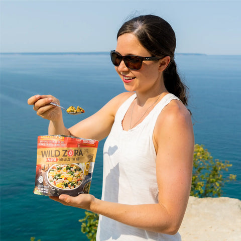 A smiling woman in sunglasses stands by a scenic blue lake, holding a spoon and a pouch of Wild Zora Meals to Go - Caldera Chicken Curry, ready to eat with greenery in the background.