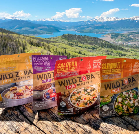 Seven Wild Zora Meals to Go - All Flavors Variety 7-Pack pouches are displayed outdoors on a wooden surface, with scenic mountains, a forest, a lake, and blue sky in the background—perfect for your next adventure!.