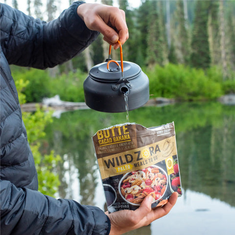 A person pours hot water from a black kettle into a Wild Zora Meals to Go - Butte Cacao Banana (Vegan) packet, which has no added sugar, with a lake and green trees in the background.