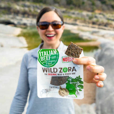 A smiling woman wearing sunglasses holds up a Wild Zora Bars - All Flavors Variety 5-Pack outdoors, with rocks and nature softly blurred in the background.