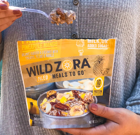 A person holds a fork with food and a yellow Wild Zora Meals to Go - Palisade Pineapple Mango (Vegan) package, a convenient single-serve meal showing the dish and product details on the front.