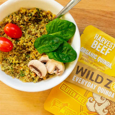 A Mediterranean quinoa bowl with cherry tomatoes, spinach, and mushrooms sits next to a Wild Zora Quinoa Meals – Harvest Beef With Mushrooms, Garlic & Oregano packet on a wooden surface.
