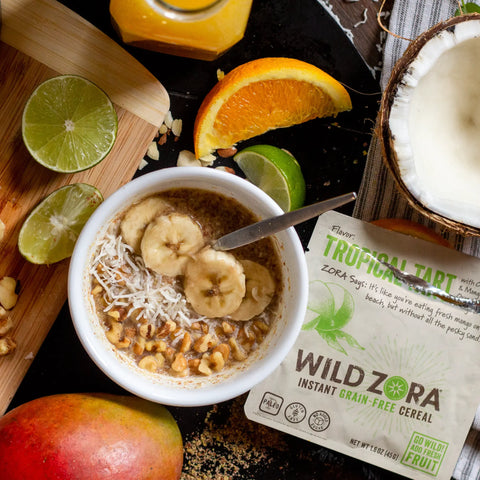 A bowl of Wild Zora Instant Cereal - Tropical Tart With Mango, Pecans, Orange & Lime sits next to its 10-pack, surrounded by fresh mango, lime, orange slices, coconut, nuts, and organic flaxseed on a rustic surface.