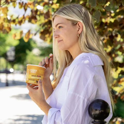 A woman with long blonde hair in a white shirt holds a cup labeled Wild Zora Vermicelli Chicken Noodle Soup (4-Pack) and a spoon, standing outdoors by a leafy tree on a sunny day.