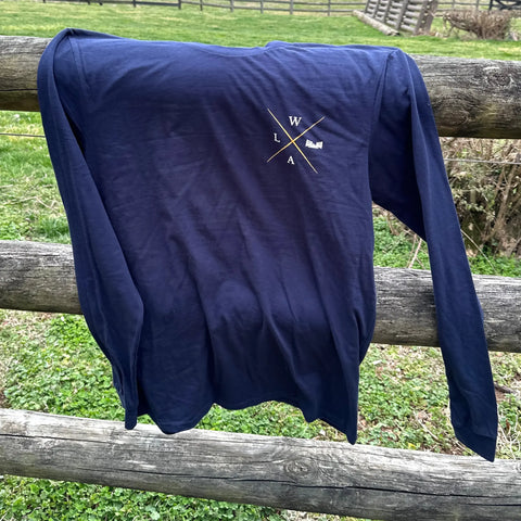 The Wilson Lane Apparel Graphic Tee Shirt - Long Sleeve, featuring a white crossed WLA chest design, hangs on a wooden fence in the grass. Made from super soft cotton, it offers the perfect blend of comfort and style.