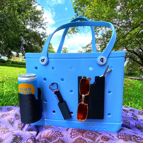 A BOGLETS Wine & Bottle Opener Charm Accessory by Chatelet Manufacturing, compatible with Bogg Bags, is attached to a light blue tote on a purple blanket outdoors, alongside High Noon and orange sunglasses under a summer sky.
