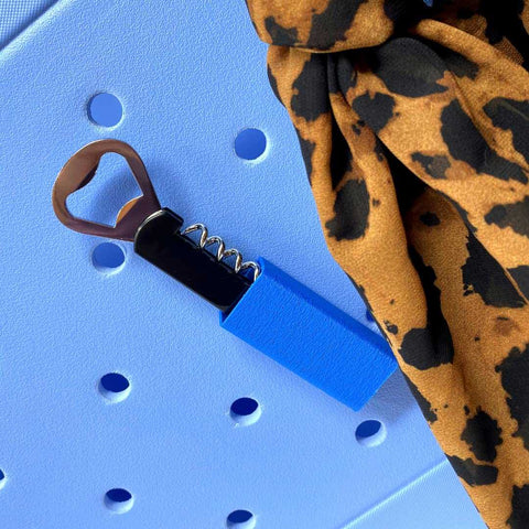 The Chatelet Manufacturing BOGLETS Wine & Bottle Opener Charm, compatible with Bogg Bags, hangs on a light blue pegboard beside leopard print fabric. It’s the perfect tote bag accessory to keep your wine opener handy.