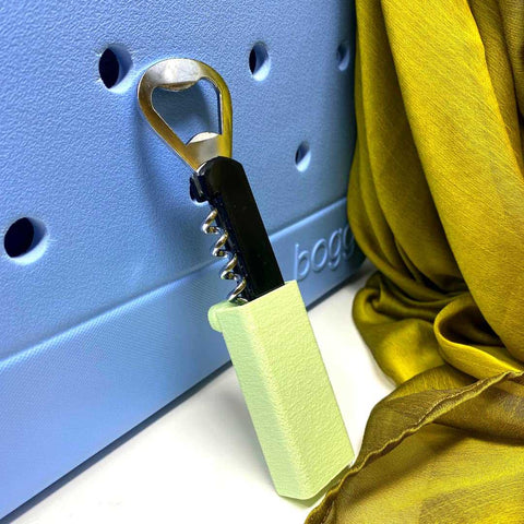 The Chatelet Manufacturing BOGLETS Wine & Bottle Opener Charm, compatible with Bogg Bags, hangs from a hole in a light blue textured surface as a handy tote accessory; a yellow-green scarf is draped nearby.