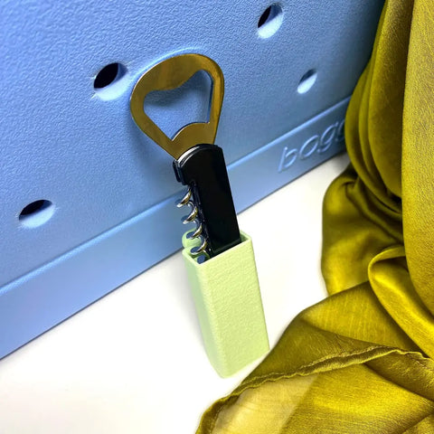 The Chatelet Manufacturing BOGLETS Wine & Bottle Opener Charm, compatible with Bogg Bags, features a light green cover and rests against a light blue tote beside yellow-green fabric—perfect for keeping your opener handy.