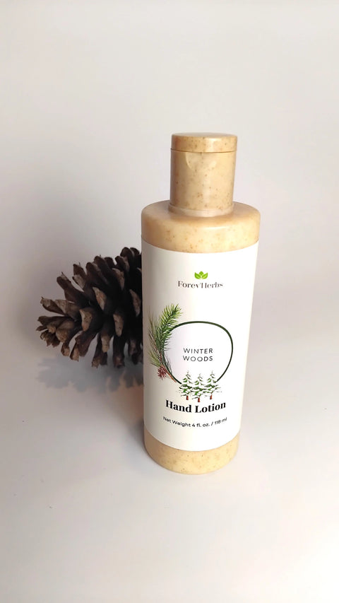 A beige Forev'Herbs Winter Woods Hand Lotion bottle stands upright on a white surface beside a pine cone. The label displays green pine branches, emphasizing its fresh pine scent and natural, soothing qualities.