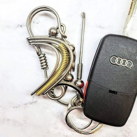 A black Audi car key with the Audi logo is attached to the Wire Coiled Birdie Keychain for Him by Alexa Martha Designs, featuring intricate silver and gold-colored metal wire sculptures that resemble puzzles or decorative keychains.