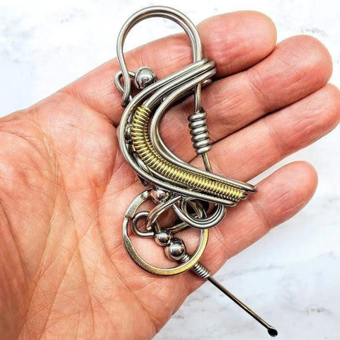 A hand holds the Wire Coiled Birdie Keychain for Him by Alexa Martha Designs, featuring handcrafted loops, coils, silver and gold tones, and seamlessly incorporated rings and beads.