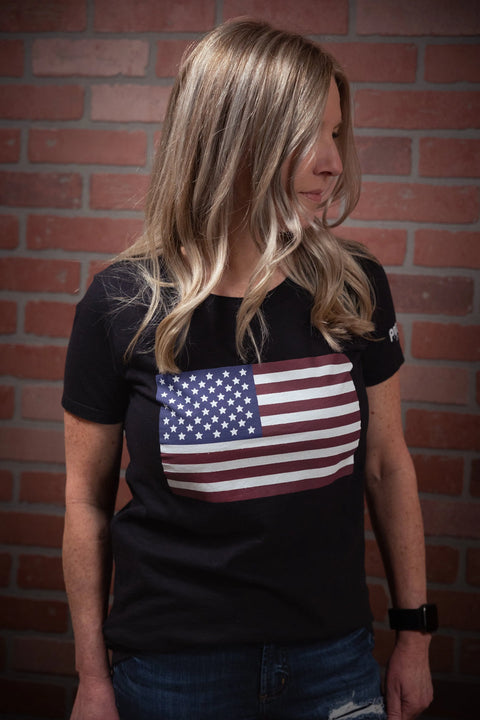 A woman with long blonde hair wears the Proudly USA American Flag - Womens Cotton T-Shirt (Black), standing by a brick wall, looking left—a stylish and patriotic choice.