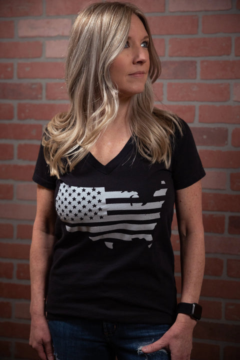 A woman with long blonde hair stands by a brick wall, wearing the Proudly USA Land That I Love Womens Cotton V-Neck T-Shirt (Black) with an America-shaped flag design and a smartwatch on her left wrist.