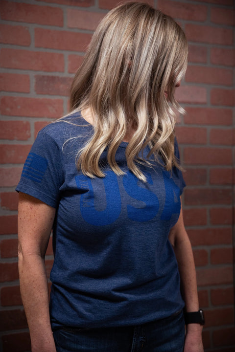 A woman with wavy blonde hair stands against a brick wall, looking down and to the side. She wears the Proudly USA - Womens TriBlend T-Shirt (Heather Denim) by Proudly USA and a smartwatch on her left wrist.
