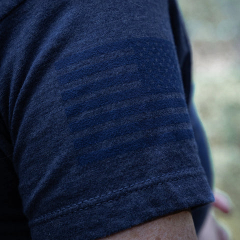 Close-up of a person’s upper arm in the Proudly USA - Womens TriBlend T-Shirt (Heather Denim) featuring a subtle, tonal American flag pattern on the sleeve. The photo is outdoors with a blurred background. Brand: Proudly USA.