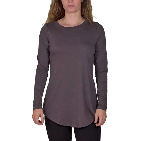 A woman with long wavy hair models the Spiritex | Sustainable Apparel Long Sleeve Tunic Tee in dark gray, paired with black pants, against a white background.