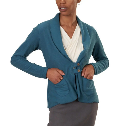 A woman stands confidently with her hands on her hips, wearing the Spiritex Lightweight Terry Tab Jacket—a teal, lightweight French terry cardigan with two pockets, shawl collar, and wooden buttons—styled over a white top and dark skirt.