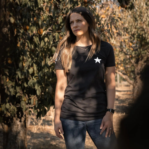 A woman stands in a sunlit orchard, wearing the Proudly USA Proudly Star Womens Cotton T-Shirt (Black), blue jeans, and a smartwatch. She looks thoughtfully to her right.