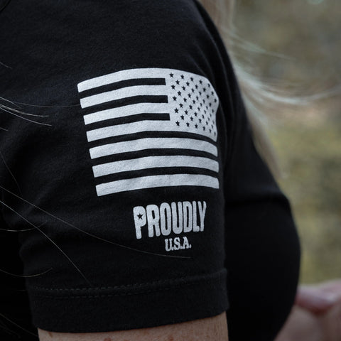 A close-up of someone wearing the Proudly Star - Womens Cotton T-Shirt (Black) by Proudly USA, made in the USA with American grown cotton, featuring a white U.S. flag and PROUDLY U.S.A. on the sleeve.
