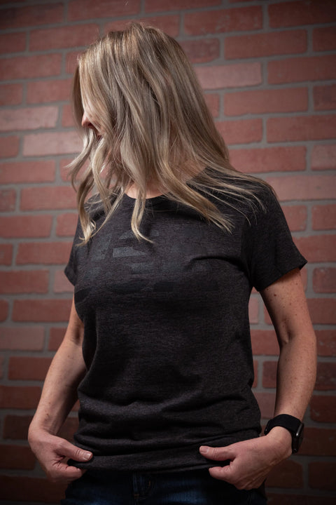 A blonde woman wears a Proudly USA Patriotic USA Womens TriBlend T-Shirt (Heather Charcoal) and jeans, standing in front of a red brick wall with her hands in her pockets, looking to her left.