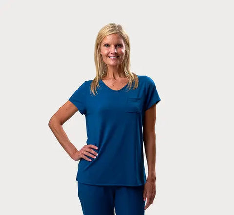 A smiling blonde woman wears the Goodnighties Short Sleeve V-Neck Tee styled as medical scrubs, posing with one hand on her hip against a plain, light background.