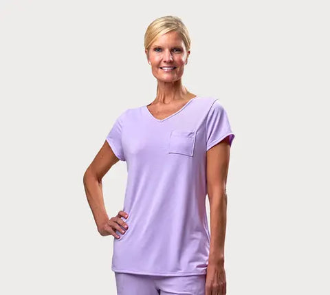 A smiling woman with short blonde hair models the Goodnighties Short Sleeve V-Neck Tee, featuring a chest pocket and matching pants, standing with one hand on her hip against a plain white background.
