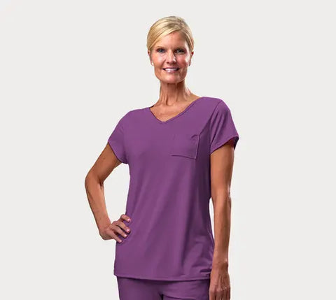 A smiling woman poses with one hand on her hip, wearing Goodnighties Short Sleeve V-Neck Tee—purple, moisture-wicking loungewear—paired with matching pants against a plain light background.