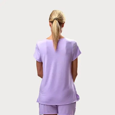 A woman with blonde hair in a ponytail stands with her back to the camera, wearing the Goodnighties Short Sleeve V-Neck Tee and matching shorts in light purple against a plain light grey background.