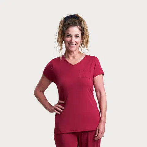 A woman with curly hair in a ponytail smiles with one hand on her hip, wearing the Goodnighties Short Sleeve V-Neck Tee and matching pants against a plain light background.