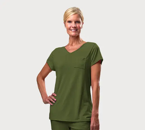 A smiling woman with short blonde hair models the Goodnighties Short Sleeve V-Neck Tee scrub top with a chest pocket and matching pants, standing with one hand on her hip against a plain white background.