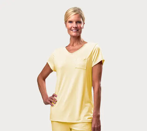 A smiling woman with short blonde hair poses with one hand on her hip, wearing Goodnighties American-made Short Sleeve V-Neck Tee and matching pants in pale yellow, set against a plain light gray background.