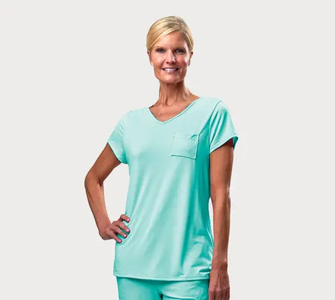 A woman with short blonde hair smiles while wearing a light turquoise Goodnighties Short Sleeve V-Neck Tee, paired with matching pants—American-made sleepwear—standing with one hand on her hip against a plain white background.