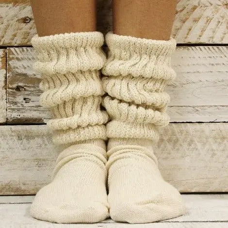 A close-up of two feet in Catherine Cole ULTIMATE Cotton Slouch Socks Women - Natural, featuring thick cream ribbed texture, shown on a rustic white wooden background. These cozy socks offer warmth and comfort to any setting.
