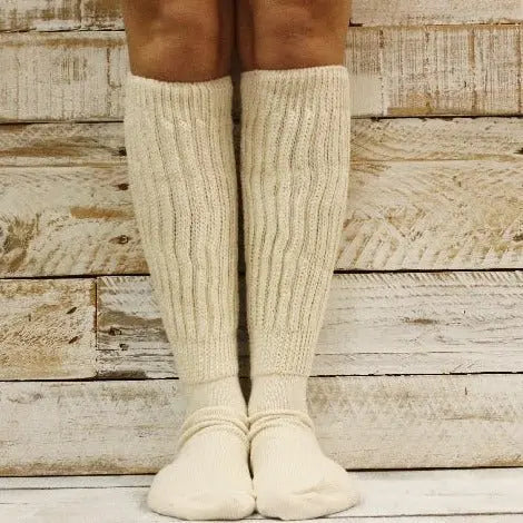 A pair of legs wears Catherine Cole ULTIMATE Cotton Slouch Socks Women - Natural, thick, cream-colored knee-highs, standing on a wooden floor with a rustic wooden wall behind.