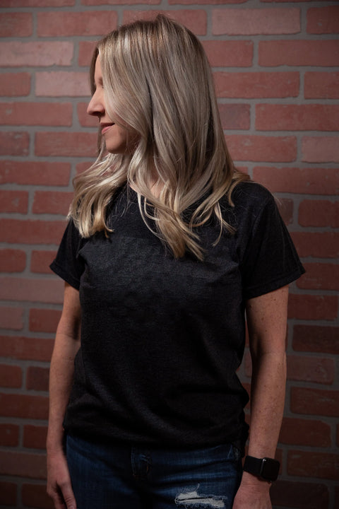 A woman with long blonde hair stands in front of a brick wall, looking left, wearing the Proudly USA Land That I Love - Womens TriBlend T-Shirt (Heather Black), ripped jeans, and a smartwatch on her left wrist.