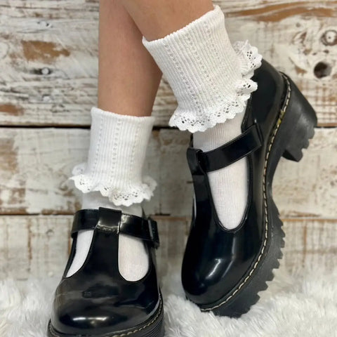 A person sports Catherine Coles SALE Lace Cuff Ankle Socks Women - White with black Mary Jane platforms, standing on a fluffy rug before a rustic wood backdrop, creating a chic vintage-inspired look.