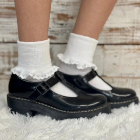 A person stands on a fluffy white rug in front of rustic wood, wearing Catherine Cole SALE Lace Cuff Ankle Socks for women in white with black Mary Jane shoes, showcasing a vintage-revival fashion look.