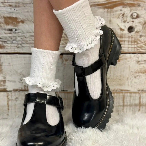 A person stands on a fluffy white rug, wearing Catherine Coles SALE Lace Cuff Ankle Socks for women in white and black T-strap Mary Jane shoes, capturing vintage-revival fashion against a rustic white wooden background.