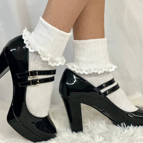 A model showcases Catherine Cole’s SALE Lace Cuff Ankle Socks for women in white, styled with glossy black Mary Jane heels on a plush white surface, capturing a vintage-revival vibe against a white background.