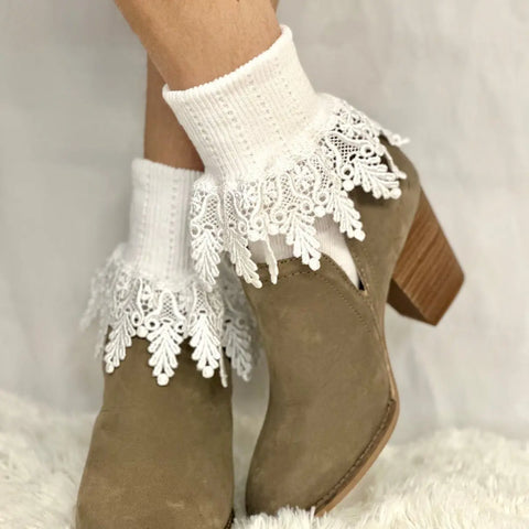 A close-up of legs wearing light brown suede ankle boots and Catherine Cole ORIGINAL Lace Cuff Socks in white, featuring decorative lace trim, set against a soft, white textured background.