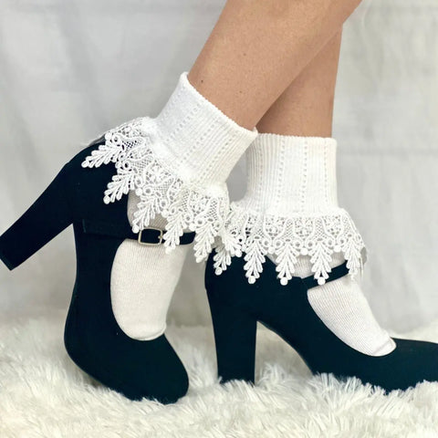 A person wears Catherine Cole ORIGINAL Lace Cuff Socks in white with black high-heeled Mary Jane shoes, standing on a fluffy white surface against a white background.