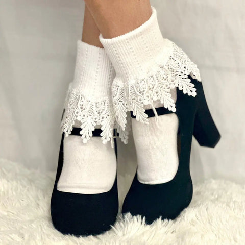 A person wears Catherine Cole ORIGINAL Lace Cuff Socks in white, paired with black Mary Jane heels, standing on a white fluffy surface against a white background.