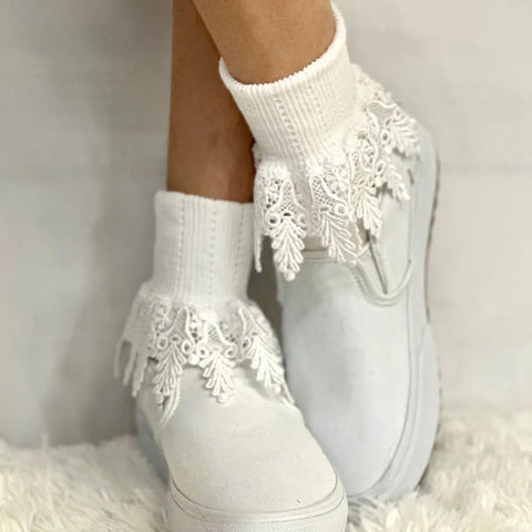 A person stands on a soft, textured white surface wearing Catherine Cole ORIGINAL Lace Cuff Socks - White with decorative trim and white slip-on shoes.