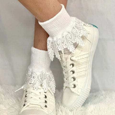 A person wears Catherine Cole ORIGINAL Lace Cuff Socks - White with white sneakers. The socks feature decorative trim around the ankles, and the background is a soft, textured white surface.