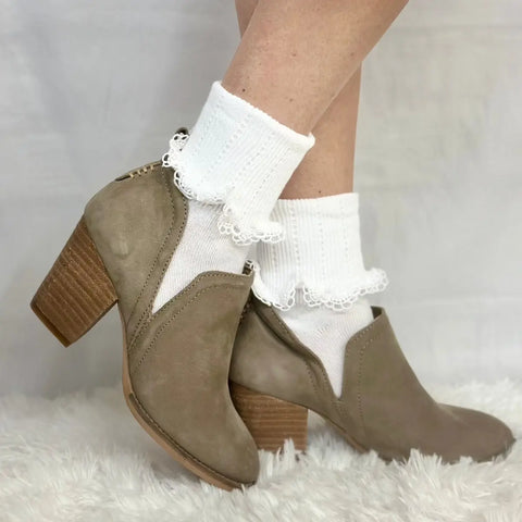 A person wears Catherine Cole SCALLOP Cuff Ankle Socks for women in white, paired with taupe suede heeled ankle boots, standing on a soft white textured surface with a plain light background.