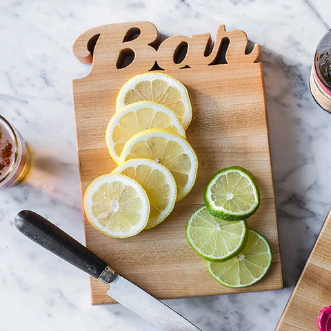 A USA made bar cutting board with lemons, limes and a knife on it lies on a marble table.
