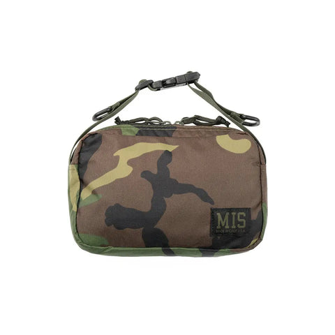 The MIS AW Shoulder Bag S in Woodland Camo is a small rectangular nylon pouch with a front zipper, top handle, and features a black MIS patch at the lower right corner.